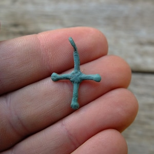 May include: A small, turquoise cross with a curved top. The cross has four arms and appears to be made of metal. The cross is held in a person's hand.