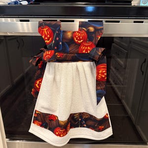 Può includere: Un canovaccio da cucina bianco con un design a tema Halloween. L'asciugamano presenta una parte superiore decorativa con un motivo di zucche arancioni, cappelli da strega e stelle su sfondo scuro. Il bordo inferiore ripete lo stesso design.