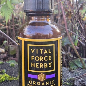 May include: A brown glass bottle with a black dropper top. The label reads "VITAL FORCE HERBS" above "ORGANIC CRYSTAL INFUSED RESTFUL SLEEP" in gold and purple lettering. The bottle is set against a natural background of greenery.