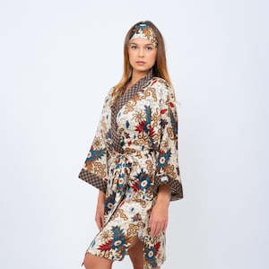 Op de afbeelding: Een crèmekleurige kimono en bijpassende hoofdband met een bloemenpatroon in rood, blauw en bruin. De kimono heeft wijde mouwen en een strikceintuur. De hoofdband past bij het ontwerp van de kimono.