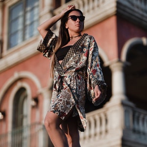 Puede incluir: Una bata estilo kimono estampada con cinturón anudado, con una mezcla de diseños florales y geométricos en tonos marrón, rojo y azul. La bata tiene mangas anchas y se lleva sobre un top negro. La persona lleva gafas de sol.