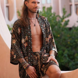May include: A black kimono-style robe and matching shorts set with a floral pattern in orange, green, and gold. The outfit is worn open, revealing the chest. The model is wearing bracelets and a necklace.