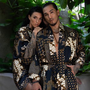 Puede incluir: Dos personas con batas azul oscuro a juego con un estampado estilo batik en blanco y marrón. Las batas tienen un diseño envolvente con cinturón. El fondo presenta un exuberante follaje verde.