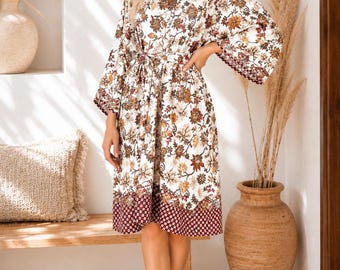 May include: A white floral print robe with a V-neck and a tie waist. The robe features a pattern of brown and orange flowers and a burgundy border. The robe is knee-length and has wide sleeves.