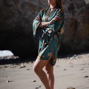 May include: A woman wearing a blue and floral patterned robe with a black sash. She is standing on a sandy beach with rocks in the background.