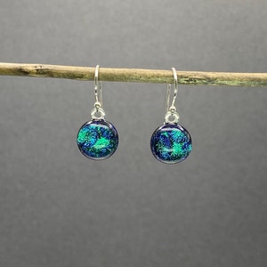 May include: A pair of round dangle earrings with a dichroic glass design. The earrings feature a dark blue background with vibrant green and blue accents. The earrings are suspended from silver-colored hooks.