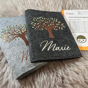 Peut inclure: Deux dossiers en feutre gris avec un motif d'arbre et les noms "Marie" et "M" imprimés dessus. Les dossiers sont sur une surface grise et duveteuse. Un dossier jaune et orange avec le texte "Kinderuntersuchungsheft" est en arrière-plan.