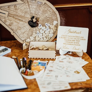 Könnte beinhalten: Ein hölzernes Gästebuch in Form des Millennium Falken mit einem herzförmigen Ausschnitt. Kleine Holzherzen sind um das Schiff verstreut. Ein Schild mit der Aufschrift "Gästebuch" und Anweisungen. Stifte, eine Schachtel mit Herzen und Schreibwaren auf dem Tisch.