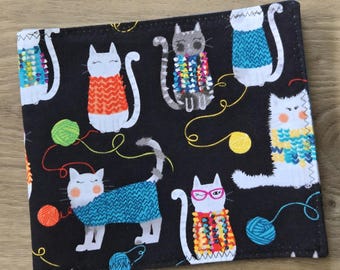 May include: A square fabric piece featuring a black background with a repeating pattern of white and gray cats wearing colorful knitted sweaters. The cats are depicted with various yarn balls in green, yellow, orange, and blue scattered throughout.
