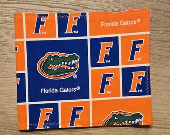 May include: A square fabric coaster featuring a repeating pattern of the Florida Gators logo, which includes a green alligator head on an orange oval, and the letter F in blue and orange. The text Florida Gators is printed in white.