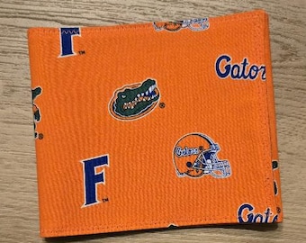 May include: A bright orange fabric wallet featuring a repeating pattern of the Florida Gators logo, a blue letter F, a football helmet with the word Gators, and the text Gators in blue. The wallet has visible stitching along the edges.