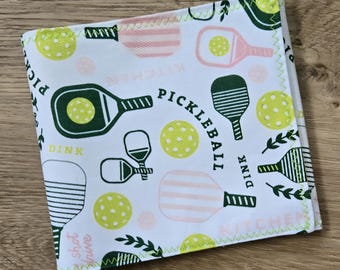 May include: A square fabric cloth featuring a white background with a repeating pattern of pickleball paddles in dark green and light pink, yellow pickleball balls, and the text "PICKLEBALL", "DINK", and "KITCHEN" in dark green and light pink.