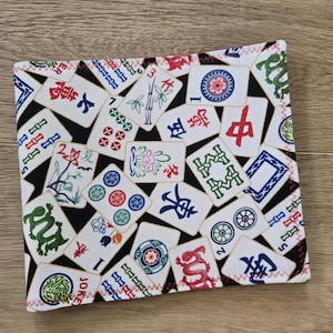 May include: A rectangular fabric item with a black background and a pattern of white Mahjong tiles. The tiles feature various symbols, numbers, and Chinese characters in red, green, and blue. The edges are finished with red zigzag stitching.