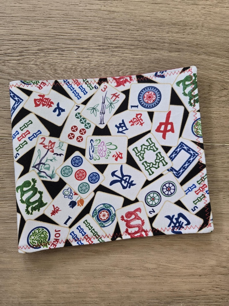 May include: A rectangular fabric item with a black background and a pattern of white Mahjong tiles. The tiles feature various symbols, numbers, and Chinese characters in red, green, and blue. The edges are finished with red zigzag stitching.