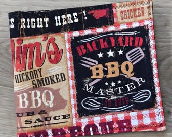 May include: A square fabric napkin featuring a collage of barbecue-themed graphics in red, black, and white. Text includes "BACKYARD BBQ MASTER," "HICKORY SMOKED BBQ," and "CHICKEN." The design incorporates a red and white checkered border pattern.
