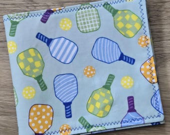 May include: A square fabric cloth featuring a light blue background with a repeating pattern of pickleball paddles and balls. The paddles are decorated with yellow and green checkered, blue striped, or blue dotted designs. The edges have blue stitching.
