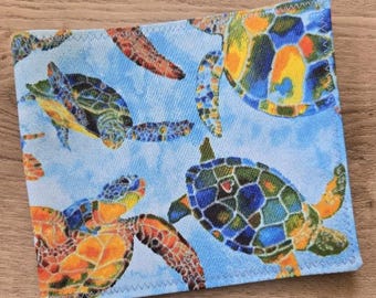 May include: A square fabric coaster featuring a vibrant pattern of sea turtles in shades of green, orange, and blue against a light blue background. The edges are finished with a decorative zigzag stitch, creating a textured and durable appearance.
