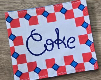 May include: A square fabric coaster featuring a white center with the word Coke embroidered in dark blue script. The border consists of a red and white checkered pattern accented with small blue diamond shapes at the corners of the squares.