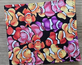 May include: A square fabric piece featuring a repeating pattern of clownfish in various shades of orange, pink, and purple against a black background. The edges are finished with a visible zigzag stitch, creating a distinct border on the textile.