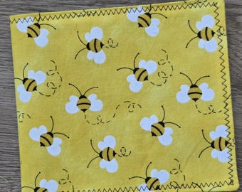 May include: A square fabric cloth featuring a repeating pattern of yellow and black bees with white wings on a mustard yellow background. The edges of the cloth are finished with a dark zigzag stitch pattern along all four sides of the square.