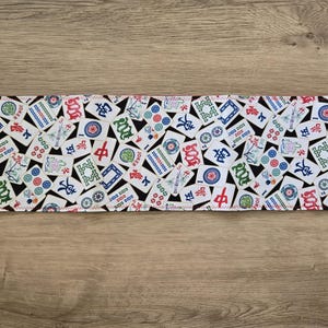 May include: Rectangular fabric with a white background covered in colorful Mahjong tiles. The tiles feature various symbols, characters, and designs in red, green, blue, and black. The edges are finished with a white zigzag stitch.