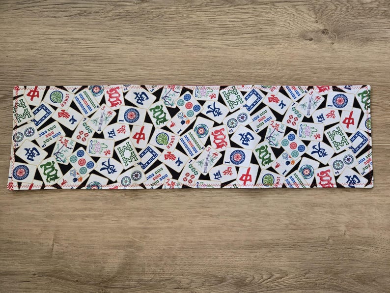 May include: Rectangular fabric with a white background covered in colorful Mahjong tiles. The tiles feature various symbols, characters, and designs in red, green, blue, and black. The edges are finished with a white zigzag stitch.