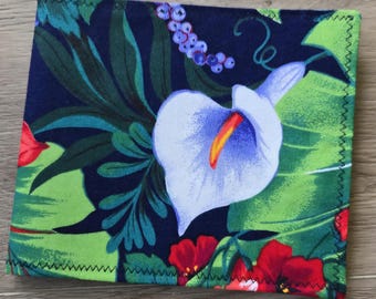 May include: A square fabric coaster featuring a dark blue background with a vibrant tropical print. The design includes large white calla lilies with orange centers, bright green monstera leaves, and small clusters of red flowers and blue berries.