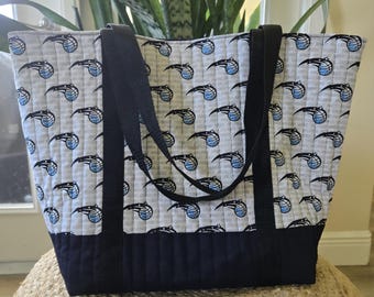 May include: A quilted tote bag featuring a repeating pattern of the Orlando Magic basketball logo on a light gray background. The bag has a solid dark navy blue bottom panel and two matching dark navy blue fabric shoulder straps.