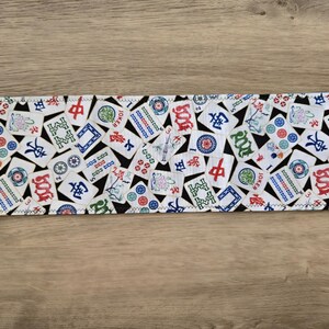 May include: A rectangular fabric item featuring a repeating pattern of colorful Mahjong tiles on a white background. The tiles have red, green, blue, and black accents. The edges are finished with a zigzag stitch.