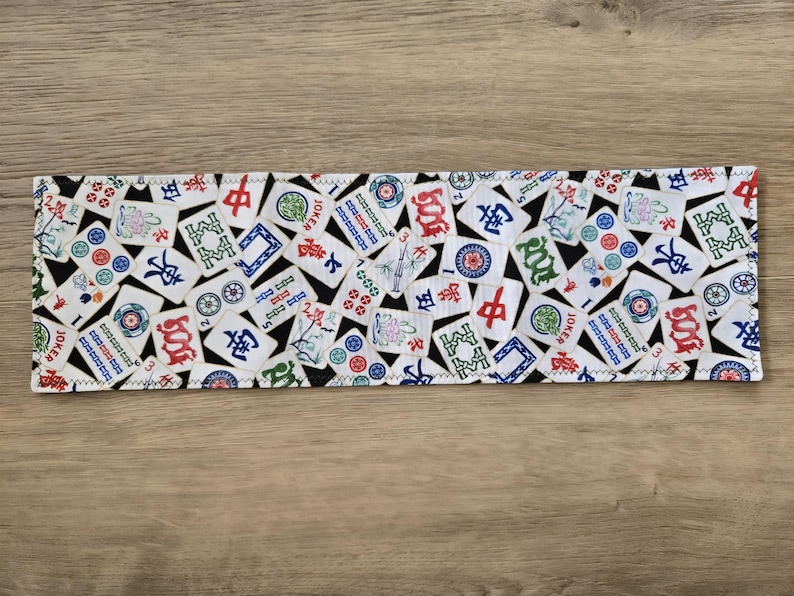 May include: A rectangular fabric item featuring a repeating pattern of colorful Mahjong tiles on a white background. The tiles have red, green, blue, and black accents. The edges are finished with a zigzag stitch.