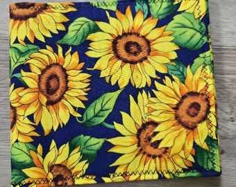 May include: A square fabric item featuring a vibrant sunflower pattern with large yellow petals and brown centers set against a deep navy blue background, accented by green leaves and finished with a black zigzag stitch along the edges.