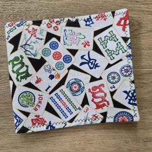 May include: A white Mahjong wallet with a black background and colorful tiles. The wallet features various Mahjong tile designs, including dragons, flowers, and numbers. The edges are stitched with white thread.