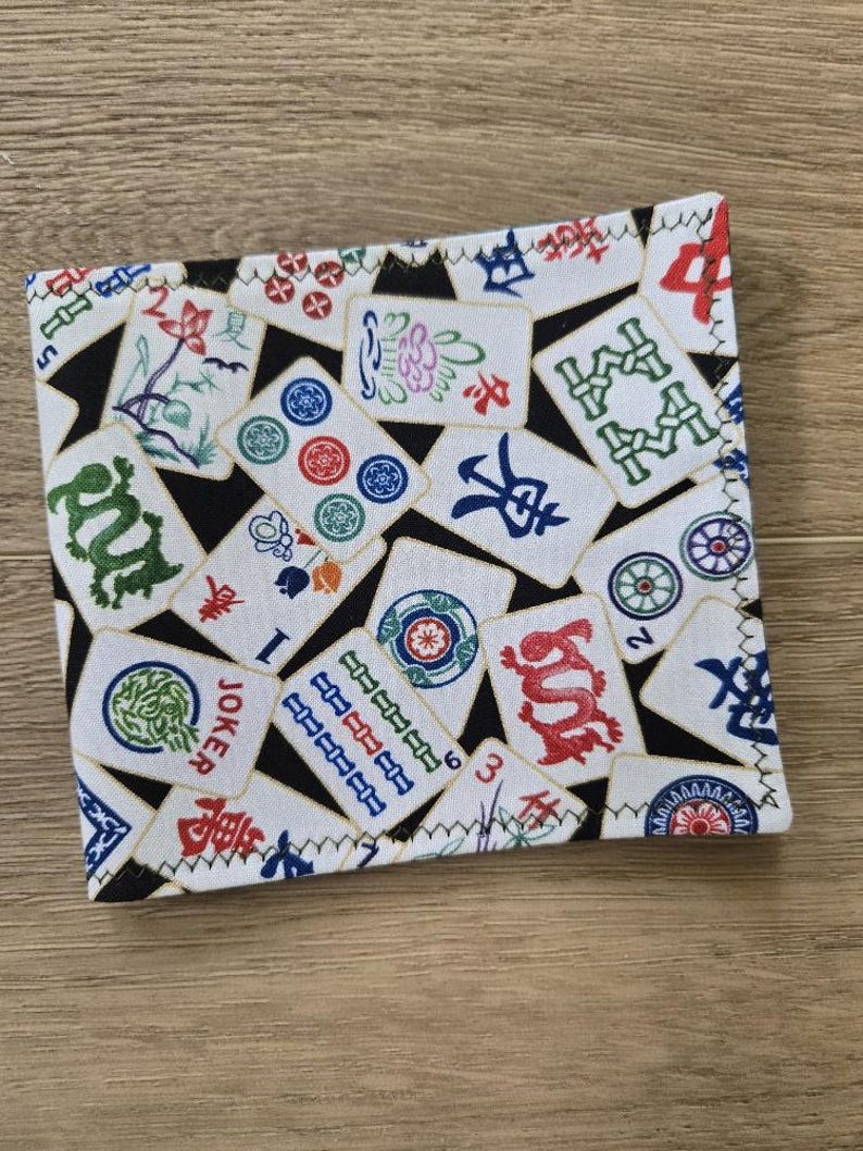 May include: A white Mahjong wallet with a black background and colorful tiles. The wallet features various Mahjong tile designs, including dragons, flowers, and numbers. The edges are stitched with white thread.