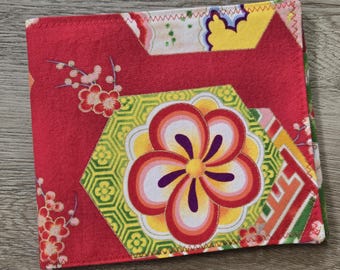 May include: A square fabric coaster featuring a vibrant red background with a central hexagonal pattern containing a stylized multi-colored flower in shades of yellow, orange, pink, and purple. The design includes small red floral sprigs.