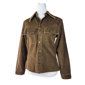 May include: A brown corduroy jacket with a button-up front, two chest pockets, and a collar. The jacket has long sleeves and silver-colored buttons. The fabric has a ribbed texture.