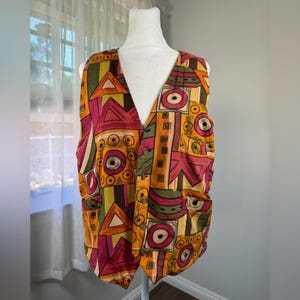 Può includere: Un gilet fantasia con scollo a V. Il gilet presenta un motivo geometrico nelle tonalità dell'arancione, del rosa, del verde e del giallo. Ha due tasche frontali ed è esposto su un manichino.