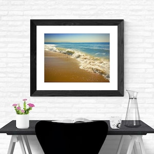 May include: A framed photograph of a wave crashing on a sandy beach. The water is a light blue and the sand is a golden brown. The sky is a light blue with some clouds.