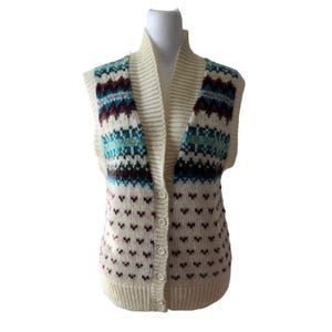 May include: A cream-colored knit vest featuring a shawl collar and a button-front closure. The garment displays a multi-colored Fair Isle pattern across the upper chest and shoulders, with small maroon heart-shaped motifs throughout the lower body.