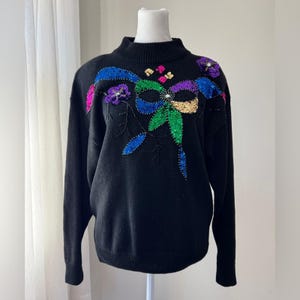 Pode incluir: Camisola preta com gola alta. A camisola é decorada com um laço de lantejoulas coloridas e um desenho floral. O laço apresenta lantejoulas azuis, verdes, douradas, roxas e rosas. A camisola é exibida em um manequim branco.