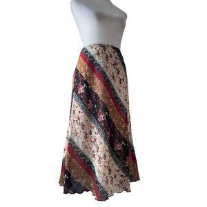 May include: A long, flowing skirt featuring a diagonal patchwork design. The pattern consists of alternating panels of cream fabric with small red and brown floral prints, solid red bands, black floral sections, and textured brown stripes.