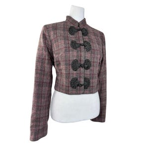 May include: A cropped jacket in a mauve and grey plaid pattern featuring a high mandarin collar and four black decorative frog closures down the front. The long-sleeved garment has a structured fit and is displayed on a white mannequin torso.