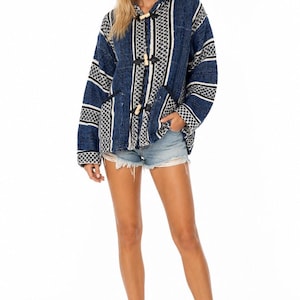 May include: A long-sleeve hooded pullover jacket in shades of navy blue, white, and black with a woven textured pattern. The jacket features a front opening with three wooden toggle closures and two side pockets, paired with light blue denim shorts.
