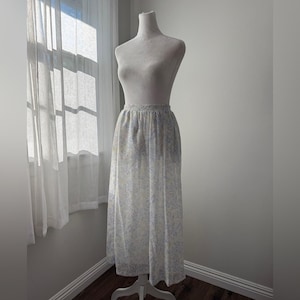 May include: A long, flowing skirt on a mannequin. The skirt is white with a delicate floral pattern in shades of blue and yellow. The skirt has a gathered waist and falls to the floor.