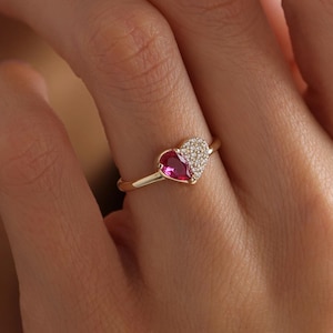 Peut inclure: Une bague en or avec un motif en forme de cœur. Une moitié du cœur est une pierre précieuse rose vif, l'autre moitié est ornée de petites pierres blanches scintillantes. La bague est portée au doigt.