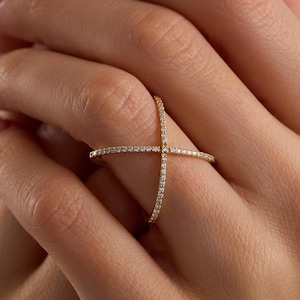 May include: A gold ring featuring a cross design, set with small, sparkling stones. The ring is worn on a finger, with the hands clasped. The ring's design is a contemporary take on a classic style, suitable for everyday wear.