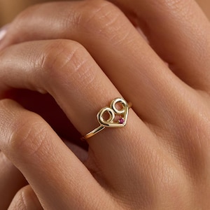 May include: A gold ring with a heart-shaped design and a small, red gemstone. The ring is worn on a finger. The ring's design includes two circles at the top of the heart. The ring is a piece of jewelry.