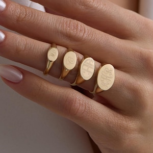 May include: Five gold signet rings of varying sizes are displayed on a hand. Each ring features an oval face with engraved designs. The rings are a classic piece of jewellery, with the largest ring on the little finger.