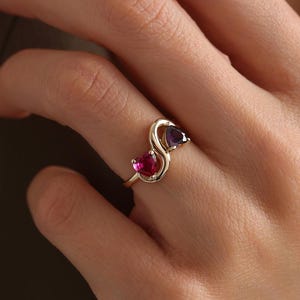 Könnte beinhalten: Ein goldfarbener Ring mit einem stilisierten Design, das zwei Edelsteine ​​trägt. Einer der Edelsteine ​​ist leuchtend rot, der andere tiefviolett. Das Design des Rings ist eine durchgehende, fließende Form.