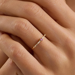 Puede incluir: Un delicado anillo de oro con una pequeña gema rosa redonda y diminutas piedras blancas. El anillo está en el dedo de una persona, con la mano enfocada contra un fondo borroso. La banda del anillo tiene un diseño texturizado.