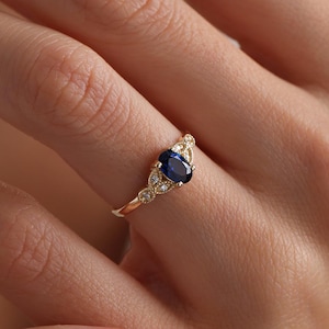 May include: A gold ring featuring a dark blue oval gemstone as the centerpiece, flanked by smaller, clear stones. The ring is set on a woman's finger, showcasing its delicate design and craftsmanship. The ring's band is thin and elegant.